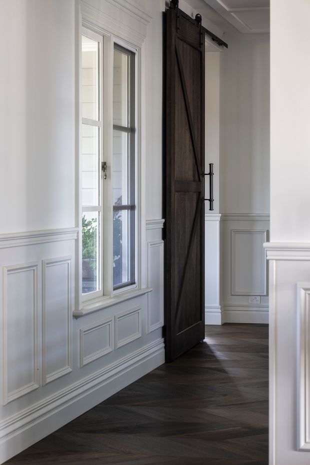 A refined interior hallway or entry area with white-painted decorative wall panelling, ornate trim, a tall multi-pane window, and a dark timber barn-style sliding door with black metal hardware. The floor is dark herringbone timber, creating contrast with the light walls and traditional detailing.