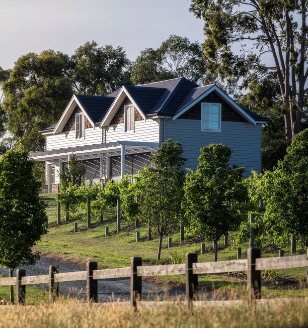 Exterior view of a light-coloured, multi-gabled house set within a landscaped rural garden or orchard. The building features horizontal weatherboard cladding, dark pitched roofs, white-trimmed dormer-like gables, and a covered verandah or pergola. A timber post-and-rail fence and rows of small trees occupy the foreground, with mature trees surrounding the property.