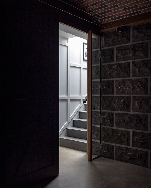 A dark, moody interior passage or stair landing viewed through an open black door. The space features a rough exposed dark stone wall framed with metal or timber grid divisions, a brick-lined ceiling, and a concrete floor. Beyond the doorway, a bright stairwell has grey concrete steps and white geometric wall panelling, creating a strong contrast between the shadowed foreground and illuminated stairs.