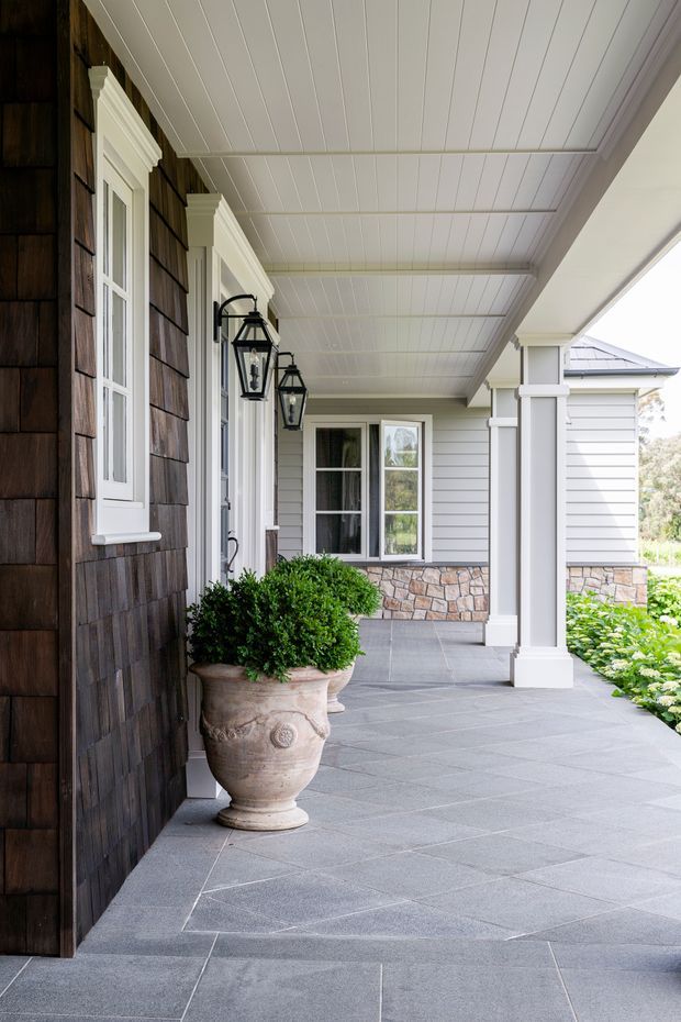 Covered front verandah along a house exterior, featuring dark timber shingle cladding, white-framed windows, decorative lantern wall lights, square white columns, a white beadboard-style ceiling, and large grey tiled paving. A stone-clad lower wall and landscaped garden edge are visible in the background.