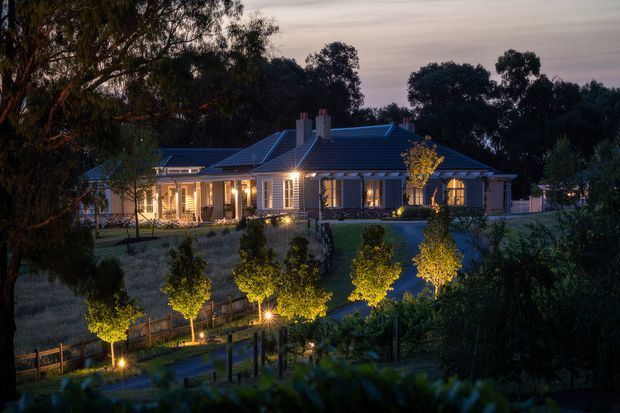 Twilight exterior view of a sprawling single-storey timber-clad residence set within a landscaped rural garden. The home features a long verandah, repeated classical-style columns, multi-pane windows with shutters, a complex hipped and gabled roofline, and warmly illuminated interiors and garden paths. Mature trees and formal planting surround the property.