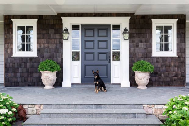 Symmetrical front entrance of a traditional-style home, featuring a dark shingle-clad facade, white-trimmed multi-pane windows, a paneled navy-blue door with sidelights, exterior lanterns, stone paving and steps, and large classical planters. The covered porch and landscaped garden edges frame the entry, with a dog seated centrally on the steps.