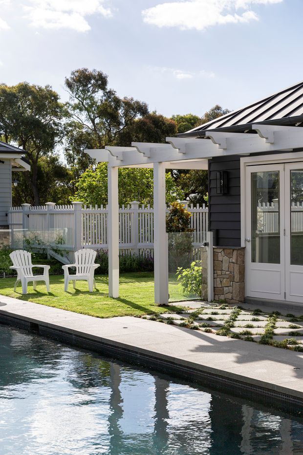 A bright landscaped residential poolside courtyard beside a dark-clad house. A white timber pergola with repeated decorative beams shades a lawn furnished with two white Adirondack chairs. A white picket fence, stone-clad wall base, glazed double doors, patterned stepping-stone paving, and the edge of a swimming pool are visible. The setting feels clean, relaxed, and traditional-coastal, with mature trees beyond the fence.