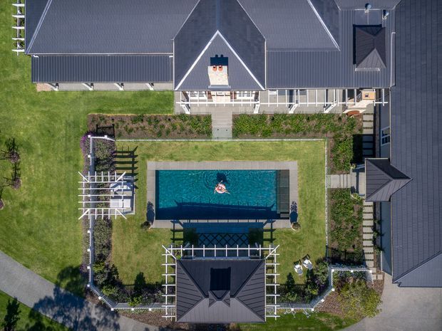 Aerial overhead view of a large residential property arranged around a landscaped central courtyard. The dark grey pitched-roof house surrounds a rectangular swimming pool, with manicured lawns, garden beds, white pergola structures, paved paths, and a detached roofed pavilion or outbuilding.