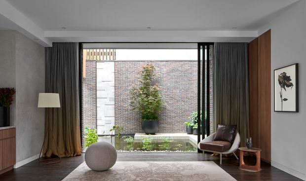A refined modern living room with dark timber flooring, a pale patterned rug, sculptural round ottoman, brown leather lounge chair, floor lamp and low timber side table. Full-height dark curtains frame large glazing looking onto a planted courtyard with a shallow water feature and brick privacy wall. A vertical timber-panelled wall and minimalist artwork add warmth and contrast.