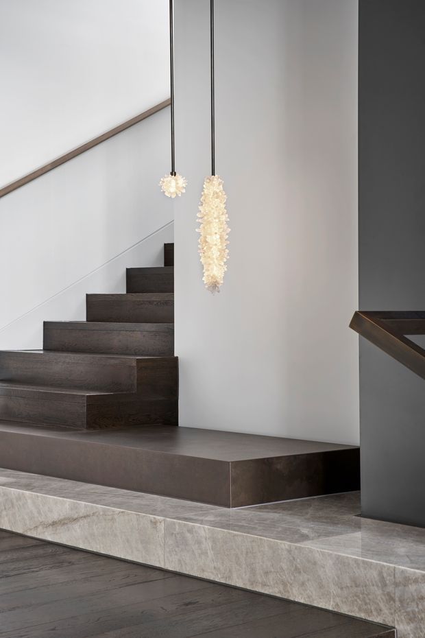 A refined modern stairway interior with dark timber floating stairs, a broad dark stone or concrete landing, pale stone-clad lower steps, smooth light walls, and two suspended sculptural pendant lights. A dark built-in cabinet or balustrade element is visible at right.