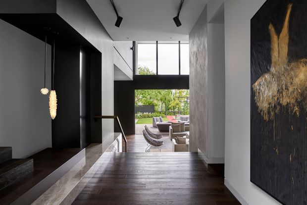 A dramatic contemporary interior hallway or entry passage with dark timber flooring, black built-in cabinetry, a stair opening, pendant lights, and tall windows overlooking a living area and garden. A large abstract artwork adds a warm gold accent against the dark wall, while white walls and high glazing balance the moody palette.