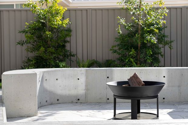 An outdoor courtyard or patio area with a minimalist concrete fire pit bench and a black freestanding fire bowl containing wood. A tall grey vertical-panel boundary fence and planted trees form the backdrop, while pale paved flooring and strong sunlight create a clean contemporary setting.