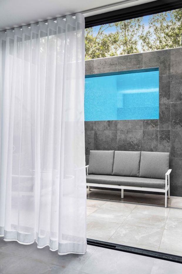 Contemporary indoor-outdoor seating area viewed through sheer white curtains and a large sliding glass opening. A grey upholstered bench sits against a dark grey tiled feature wall, with a bright turquoise rectangular pool or water feature visible above it. Large pale stone-look floor tiles and black-framed glazing create a clean, modern composition.