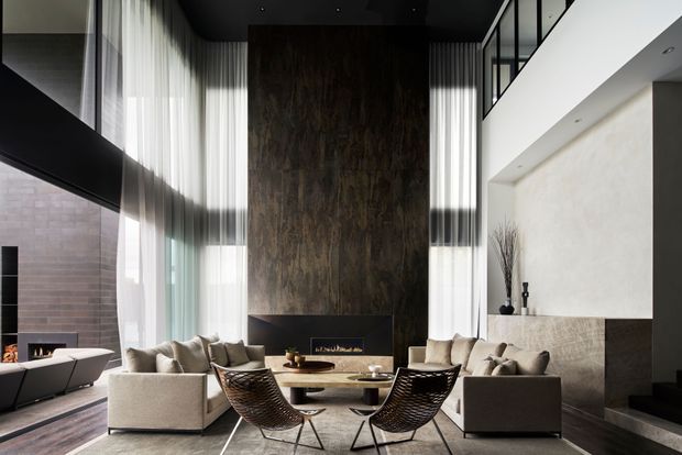 A sophisticated double-height living room with tall sheer curtains, a dramatic dark stone or timber-look feature wall surrounding a linear fireplace, and a pair of light neutral sofas facing a low coffee table. Two sculptural woven lounge chairs sit in the foreground. A mezzanine-level black-framed glazed balustrade and staircase are visible to the right, with strong natural light creating a refined, high-contrast atmosphere.