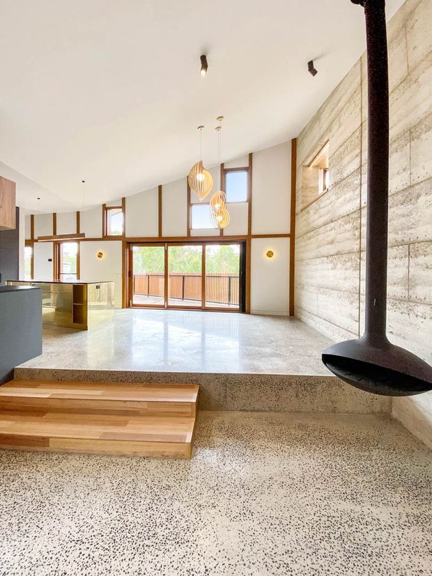 Open-plan contemporary living space with a split-level polished terrazzo floor, timber steps, exposed timber-framed glazing, and a prominent suspended black fireplace. A compact kitchen is visible to the left, while the room opens to a timber balcony or deck through large glazed doors. The space has a high sloping ceiling, warm pendant lighting, and a textured light concrete feature wall.