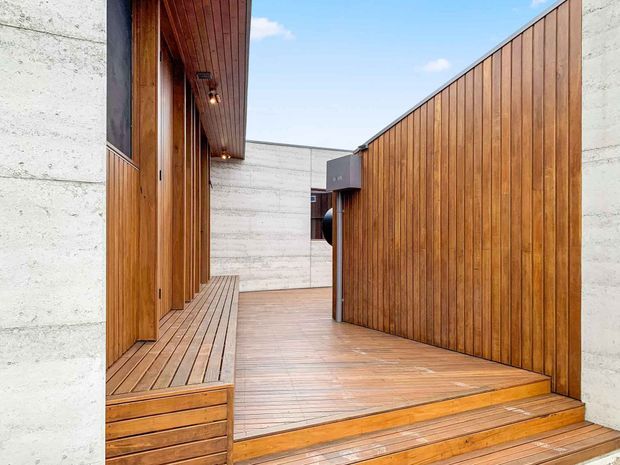 A modern timber-clad entry courtyard or walkway with warm vertical wood walls, matching timber decking and built-in bench seating. Pale exposed concrete walls frame the space, with a covered entry and recessed lighting creating a clean, contemporary architectural composition.
