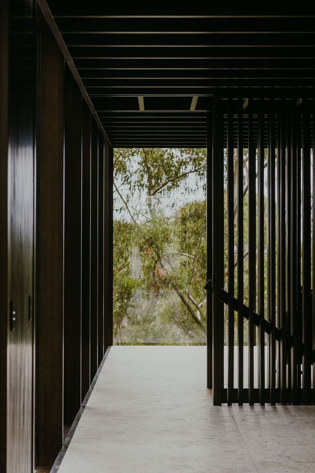 A dark, refined architectural passageway or landing with a pale concrete floor, black timber or metal vertical slats, and a strong linear ceiling structure. The corridor opens toward leafy vegetation, creating a framed connection to the outdoors. The composition emphasizes rhythm, shadow, and contrast between the dark structure and the brighter natural view.