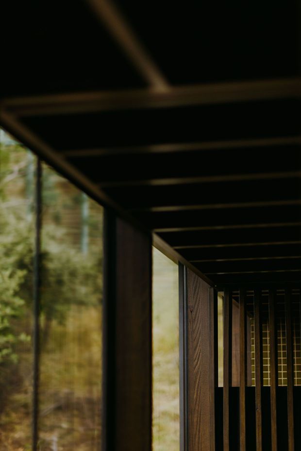 A dark, atmospheric architectural passage or corridor framed by exposed dark timber beams and vertical structural elements. Floor-to-ceiling glazing along the left side looks onto dense woodland, while timber panels and a gridded end wall create strong linear perspective and layered depth. The image is moody and minimal, with warm brown wood against near-black framing and muted green forest beyond.