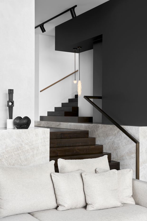 A contemporary interior featuring a dark timber staircase with a slim black handrail, rising beside a dramatic charcoal feature wall. Pale textured stone or marble-look wall surfaces contrast with the dark stair treads and cabinetry-like forms. A light upholstered sofa with cushions occupies the foreground, while minimalist black track lighting and sculptural pendant lights add to the refined monochromatic scheme.