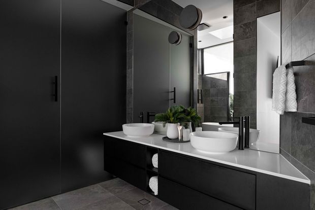 A sophisticated contemporary bathroom featuring a double vanity with white countertop and two above-counter white basins, matte black cabinetry, large-format dark grey wall and floor tiles, and a large mirrored wall. Black tapware, hardware, and framed shower or window elements reinforce the monochromatic scheme. A towel rail and small decorative plants add functional and natural accents.