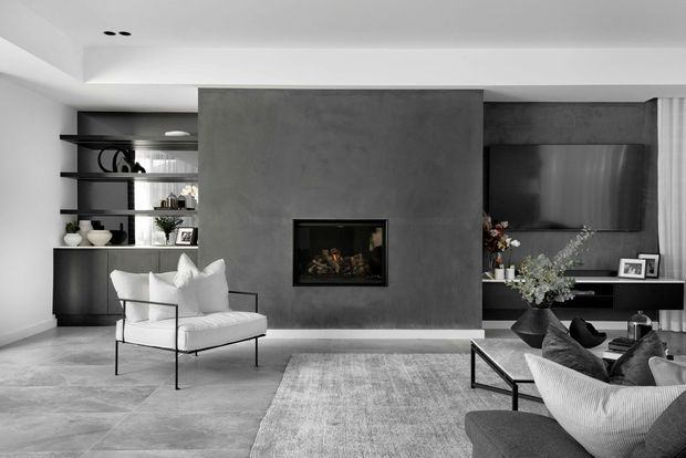 Contemporary monochromatic living room with a large dark grey feature wall incorporating a built-in fireplace, floating TV cabinetry, and open display shelving. The room features pale stone-look flooring, a neutral rug, black-framed lounge chairs, a grey sofa, and restrained decorative styling. Recessed ceiling lighting and clean-lined joinery create a refined, minimalist atmosphere.