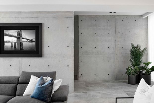 Contemporary living room with a grey sectional sofa, white and blue cushions, and a large framed monochrome photograph. The space features exposed smooth concrete walls with visible form-tie marks, a dark vertical structural or joinery element, and large grey tiled flooring. Potted greenery adds a small natural accent, while recessed ceiling lighting keeps the minimalist interior bright and restrained.