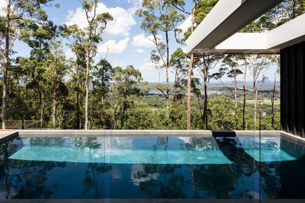 A modern elevated pool terrace overlooking a dense eucalyptus woodland and distant green valley. The rectangular swimming pool sits behind a frameless glass balustrade, with a sharply projecting flat roof and dark vertical exterior cladding framing the right side. Bright daylight, reflective water, and extensive glazing create a tranquil connection to the surrounding landscape.