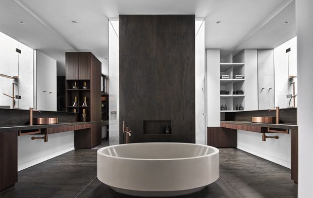 A spacious, symmetrical contemporary bathroom with a freestanding oval bathtub centred in front of a dark full-height feature wall and fireplace-like inset. The room includes twin floating timber-veneer vanities with copper-toned fixtures, large mirrors, open shelving, white cabinetry, and dark stone-look floor tiles. Recessed ceiling lighting and a restrained palette create a refined, high-contrast atmosphere.