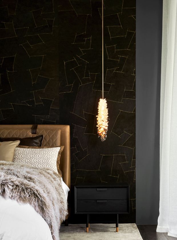 A moody, luxurious bedroom vignette featuring a black textured feature wall with irregular stitched geometric lines, a tan quilted leather bedhead, layered neutral bedding and throws, a black bedside cabinet, and a warm sculptural pendant light. Dark timber flooring, a pale rug, and sheer white curtains add contrast.