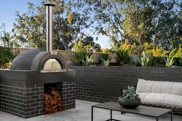 Outdoor entertaining area featuring a freestanding dark brick pizza oven with a tall metal flue, a paved patio, black brick retaining wall, raised garden beds, and a contemporary outdoor sofa and coffee table. The scene is landscaped and styled as an outdoor lounge and cooking space.