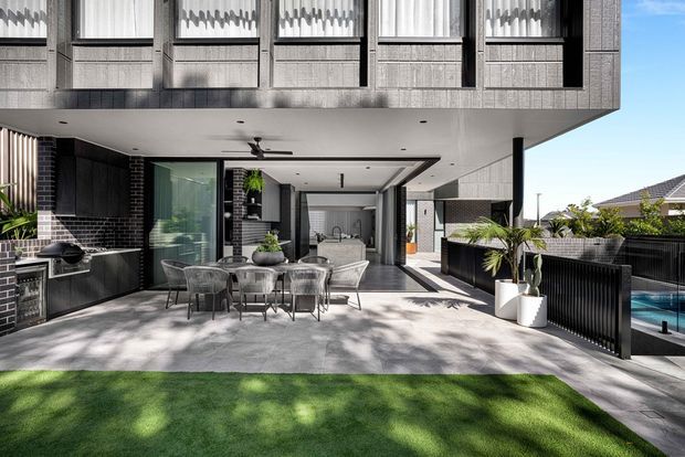 Contemporary two-storey residential courtyard entertaining area beneath a deep cantilevered upper level. The outdoor space features a concrete terrace, dining setting, built-in barbecue kitchen, dark brick and vertical ribbed metal wall finishes, black-framed glazing, screening, planters, and a swimming pool visible at the side. The upper facade has a regular grid of tall windows with patterned privacy curtains or screening, while neighbouring pitched-roof homes are visible beyond.