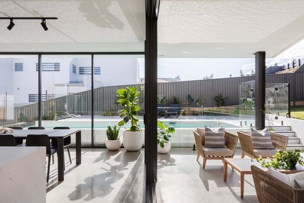 Bright, contemporary indoor-outdoor living and dining area opening through floor-to-ceiling glazing onto a swimming pool and landscaped courtyard. The space has a pale polished concrete floor, white textured ceiling, slim dark window and door frames, a dining table with dark chairs, and woven timber lounge chairs arranged around low tables. Strong daylight and the seamless connection to the pool create an airy, relaxed resort-like setting.