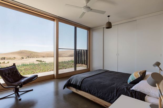 A minimalist bedroom with a low timber-framed bed, dark bedding, built-in white wardrobes, a sculptural lounge chair, pendant lighting and a ceiling fan. Expansive floor-to-ceiling glazing opens to a sparse sandy landscape and provides abundant natural light.