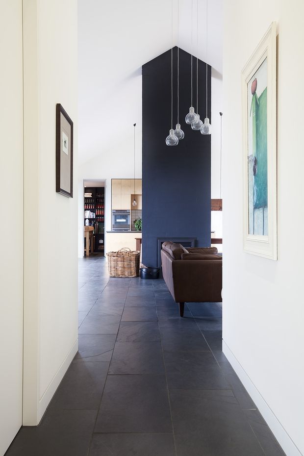 A view from a narrow hallway into an open-plan living and kitchen area. The space has dark large-format tiled flooring, white walls, a tall black feature wall or partition with hanging glass pendant lights, a brown leather sofa, light timber kitchen cabinetry, and framed artwork. The interior is bright, clean, and contemporary with strong black-and-white contrast.