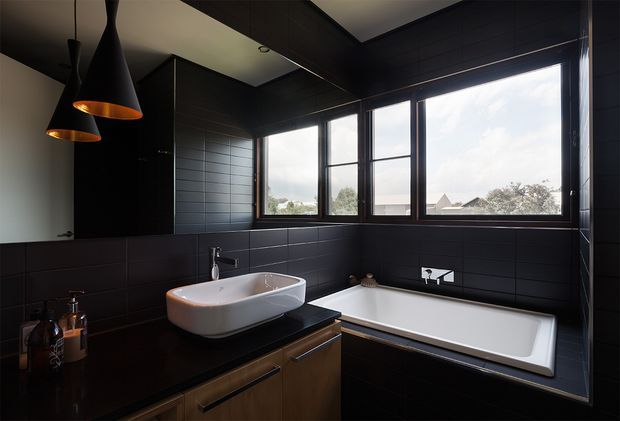Contemporary dark bathroom featuring a freestanding white vessel basin on a timber vanity, a built-in white bathtub, charcoal rectangular wall tiling, black-framed windows, dark cabinetry, and sculptural black pendant lights. Natural light enters through the large windows, creating a dramatic yet refined atmosphere.