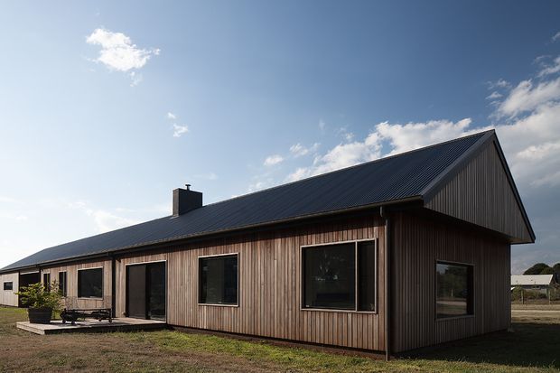 Single-storey detached house viewed from the exterior, featuring vertical timber wall cladding, a dark corrugated pitched gable roof, large dark-framed windows, and a simple linear plan. The home sits in an open rural or semi-rural setting under a partly cloudy sky.