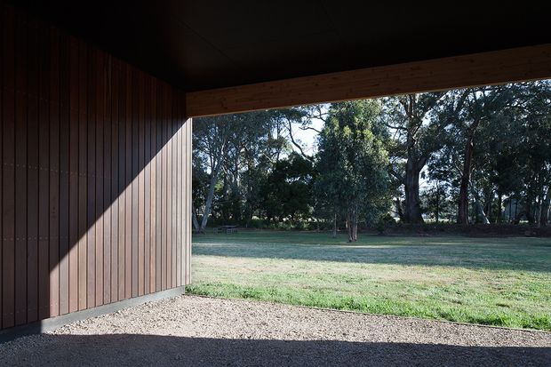 View from a covered outdoor structure through a wide rectangular opening toward a grassy, tree-lined landscape. The structure has dark timber vertical wall cladding, exposed timber framing at the opening, and a dark ceiling, creating a shaded threshold between the building and the garden.