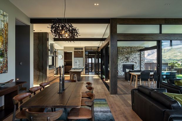 Open-plan contemporary dining and living area with a long dark timber dining table, leather dining chairs, and a sculptural branch-like pendant light. The space connects directly to a modern kitchen with island and extensive dark-framed glazing. A separate dining nook is visible through the glazing, alongside a textured stone wall and a scenic exterior backdrop. Timber flooring, dark structural beams, and black-framed window and door systems create a refined, warm, high-contrast interior.