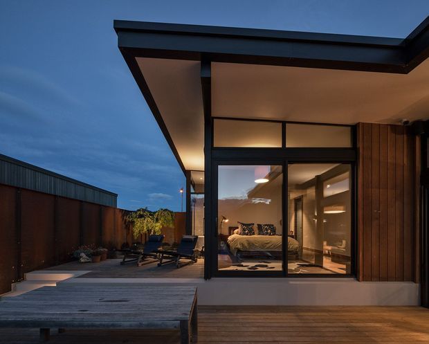 Twilight exterior view of a contemporary single-storey dwelling opening onto a timber deck and enclosed courtyard. A broad dark-framed glazed wall reveals a bedroom and warm interior lighting, while the projecting flat roof, exposed posts, vertical timber cladding, and sheltered outdoor seating define the architecture.