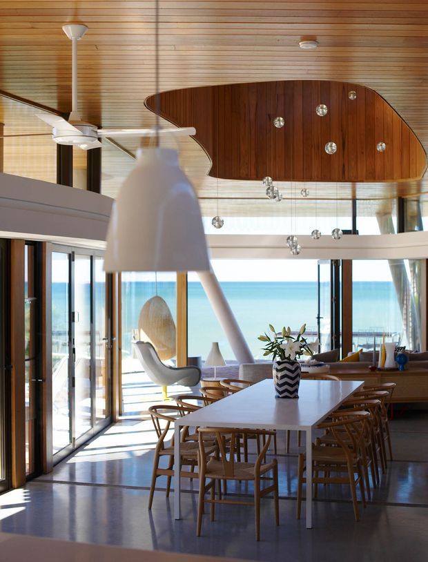 A bright, open-plan coastal dining and living space with expansive glazing overlooking the ocean. The room features a long white dining table with timber dining chairs, sculptural pendant lighting, a white ceiling fan, timber-framed sliding doors, polished concrete flooring, and a distinctive curved timber-paneled ceiling feature with small recessed lights. A lounge area and sculptural lounge chair are visible beyond the dining zone.