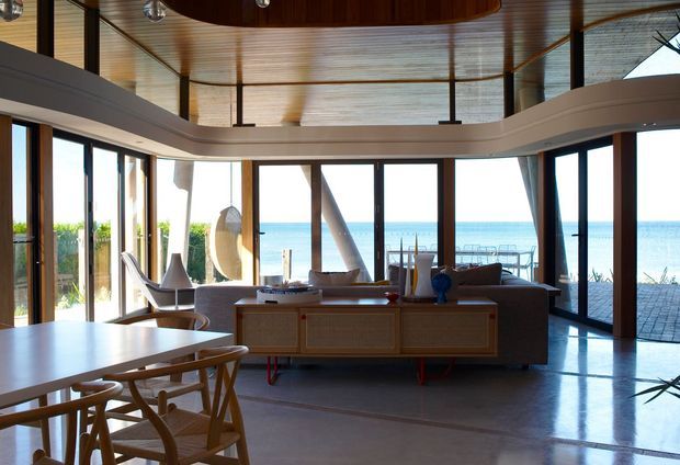 A bright, open-plan coastal living and dining area with floor-to-ceiling glazing overlooking the ocean. The room features a large curved architectural ceiling and mezzanine-like upper edge, warm timber ceiling lining and window frames, polished concrete flooring, sculptural pendant lighting, a low timber media console, and relaxed contemporary furnishings. An outdoor paved terrace is visible beyond the glazing.