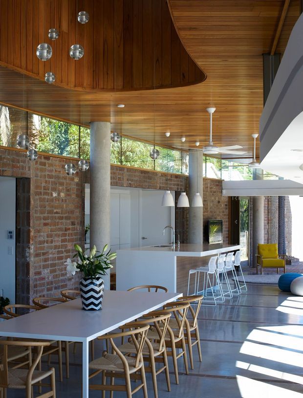 Open-plan contemporary interior combining a dining area and kitchen, with a long white dining table and timber-framed chairs in the foreground. The kitchen features a white island with integrated sink, pendant lights, and white bar stools. Exposed red brick walls, cylindrical concrete columns, expansive glazing, and a richly detailed curved timber ceiling create a warm industrial-modern character. A bright lounge area is visible beyond the kitchen.
