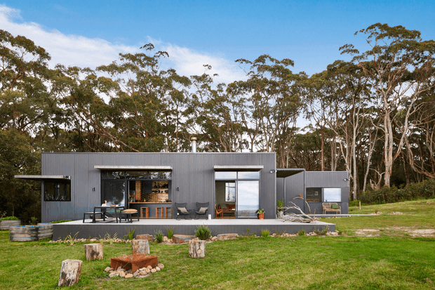 A compact single-storey contemporary dwelling set within a wooded landscape. The house has a dark grey corrugated metal-clad facade, a low simple roofline, large glazed openings, and a covered outdoor deck with seating. A separate small outbuilding or extension is visible to the right. The landscaped foreground includes lawn, tree stumps, raised garden elements, and an outdoor fire area.