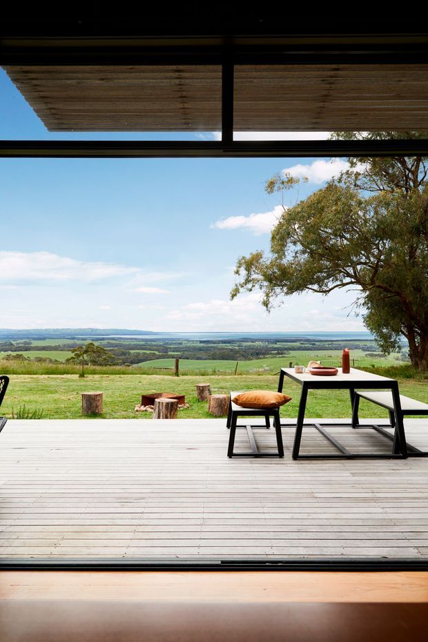 View from inside a modern glazed building onto a spacious timber deck overlooking a rural pastoral landscape and distant water or flat horizon. The deck has simple black outdoor dining furniture, stools, cushions and a small fire area with log seating. A broad timber pergola or slatted roof shades the upper glazing, while a mature tree frames the right side.