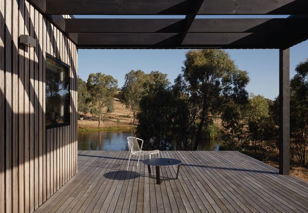 A contemporary timber-clad building opening onto a broad timber deck beside a calm waterway. A dark exposed-beam pergola provides shade over the deck, which is furnished sparsely with a small round table and wire chairs. The view looks across the water toward native vegetation and a dry, lightly wooded landscape.