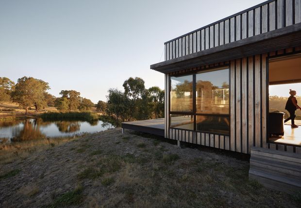 Exterior view of a compact contemporary pavilion-like house beside a calm pond, set within a natural rural landscape. The building has dark vertical timber cladding, extensive floor-to-ceiling glazing, a projecting deck, and a rooftop terrace enclosed by simple dark metal balustrades. Warm interior light and a silhouetted person are visible through the glazing.