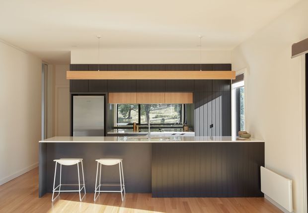 A contemporary open-plan kitchen viewed from the living area, centred on a large dark ribbed island with two white bar stools. The kitchen features dark vertical-panel cabinetry, warm timber overhead cabinetry and pendant light detailing, pale walls, timber flooring, stainless-steel appliances, and a window looking onto greenery. The space is clean, warm and moderately spacious with a standard-height ceiling.
