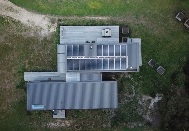 Aerial top-down view of a low, contemporary building with multiple corrugated metal roof sections. Two large arrays of solar panels dominate the central roof, with a small rooftop unit and skylight-like feature visible. The building sits within a grassy, sparsely landscaped site with outdoor furniture around it.