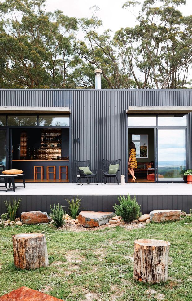 A modern single-storey pavilion-like home with a dark vertically corrugated metal facade, broad glazed openings, and a covered timber-deck terrace. One opening reveals a compact home bar with stools and shelving, while another leads into a living area. The foreground features a landscaped garden with native planting, rocks, grass, and rustic timber stump seats. A chimney rises above the roof, and a distant landscape is visible through the glazing.
