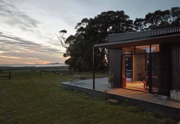 A modern single-storey rural home or retreat with dark vertical corrugated cladding, extensive glazing, and a timber-lined covered terrace. The building sits beside open pasture with mature trees and broad distant coastal or rural views at sunset. A raised deck and paved outdoor platform extend from the glazed living area.