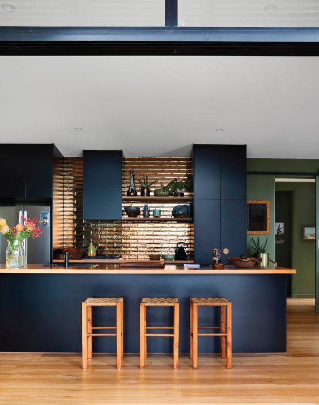 A stylish open kitchen with a dark navy island and tall cabinetry, warm timber flooring and benchtop, and a textured exposed brick splashback with open shelving. Three woven-seat timber stools line the island, while a green-painted adjoining hallway is visible to the right. The space combines strong dark cabinetry with warm natural materials and colourful decorative accents.
