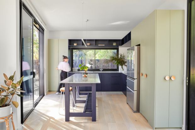 Bright contemporary kitchen with a central island, pale green full-height cabinetry, dark navy lower cabinets and overhead joinery, integrated appliances, and extensive black-framed glazing. Timber flooring and abundant natural light create an airy, refined space.