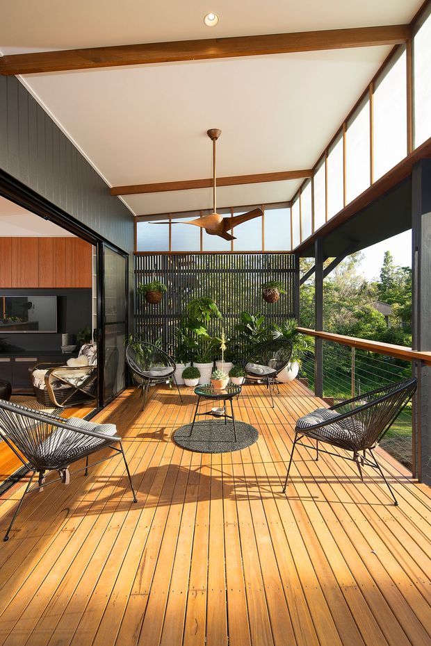 A bright, elevated timber deck or balcony adjoining a modern mid-century-style home. The space features warm timber decking, slim dark metal railings, large floor-to-ceiling glazing, exposed timber beams, a high sloping ceiling, a ceiling fan, and several black woven lounge chairs around a small round coffee table. Potted plants and a vertical slatted privacy screen create a leafy outdoor retreat, while the open glazing reveals part of the adjacent living area.