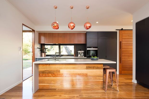 A warm, contemporary kitchen viewed straight on, centred around a large island with a timber-clad front and pale benchtop. The room features rich timber flooring, timber upper cabinetry, dark lower and full-height cabinetry, a black-framed window, and three copper-toned pendant lights. A timber pivot or sliding door and an open glazed door provide additional architectural detail and natural light.